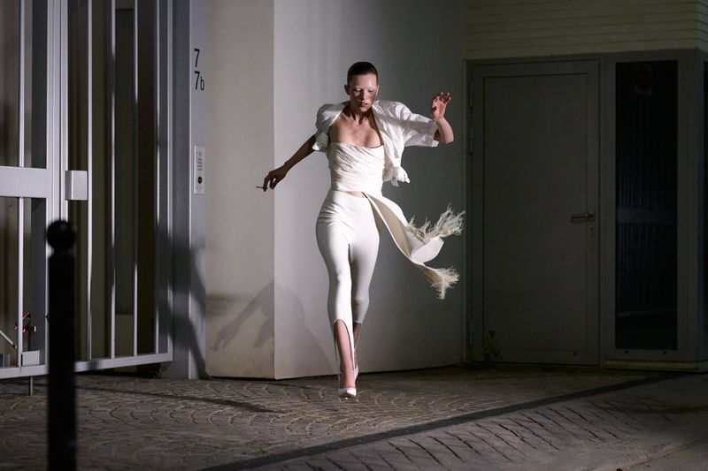 Fashion model in white outfit strikes a pose outdoors at night, wearing a jacket, top, leggings, and high heels, and holding a cigarette.