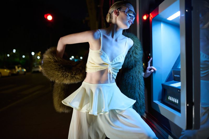 Woman in fashionable outfit stands at ATM on city street at night, illuminated by the glow of the ATM screen and nearby streetlights.