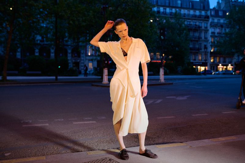 A fashionable adult female poses in an off-white dress and dark shoes in an urban setting at night, with buildings in the background.