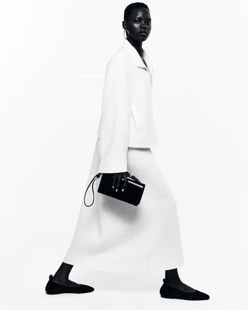 Elegant Black female fashion model in a white jacket and dress holding a black purse walks against a white background in studio lighting.