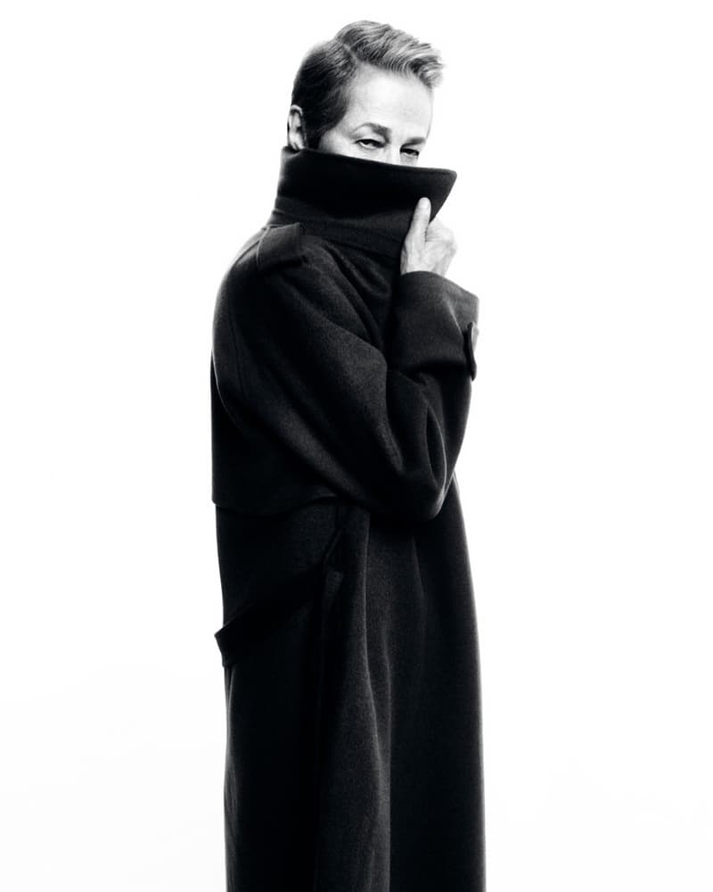 Monochromatic portrait of a person in a dark overcoat with the collar pulled up, partially obscuring their face against a white backdrop.