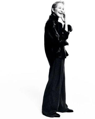 Full-length black and white portrait of a mature adult woman smiling, wearing a dark top and dark pants in a white photo studio.