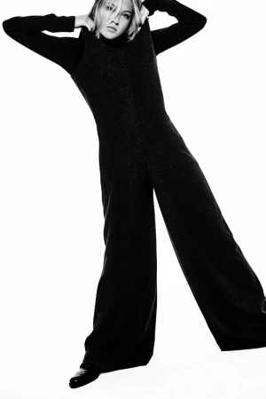 Fashion shot: woman in sleeveless jumpsuit and black long-sleeved top posing in a white studio. Black and white fashion portrait.