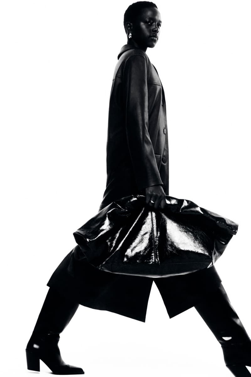 Model in dark leather coat carrying a large black handbag against a bright white background