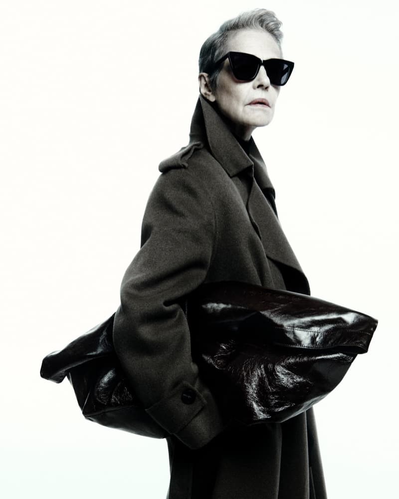 Fashionable woman in sunglasses and a coat holding a leather bag against a clean white background in a studio setting.
