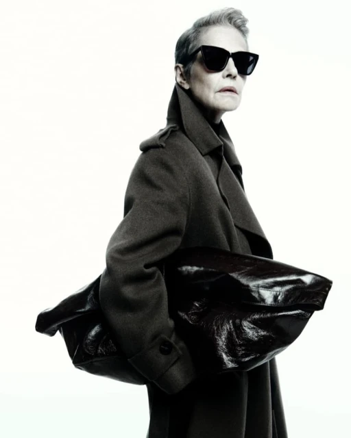 Fashionable woman in sunglasses and a coat holding a leather bag against a clean white background in a studio setting.