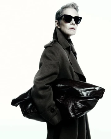 Fashionable woman in sunglasses and a coat holding a leather bag against a clean white background in a studio setting.