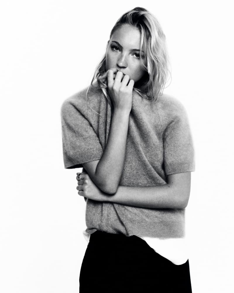 Black and white portrait of a young fair-skinned woman with blonde wavy hair, wearing a grey sweater and black skirt, indoors.