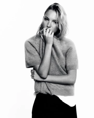 Black and white portrait of a young fair-skinned woman with blonde wavy hair, wearing a grey sweater and black skirt, indoors.