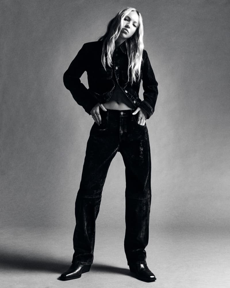 Monochrome portrait of a fair-skinned adult woman in dark denim jacket, jeans, and boots, posing in a photo studio with a grey background.
