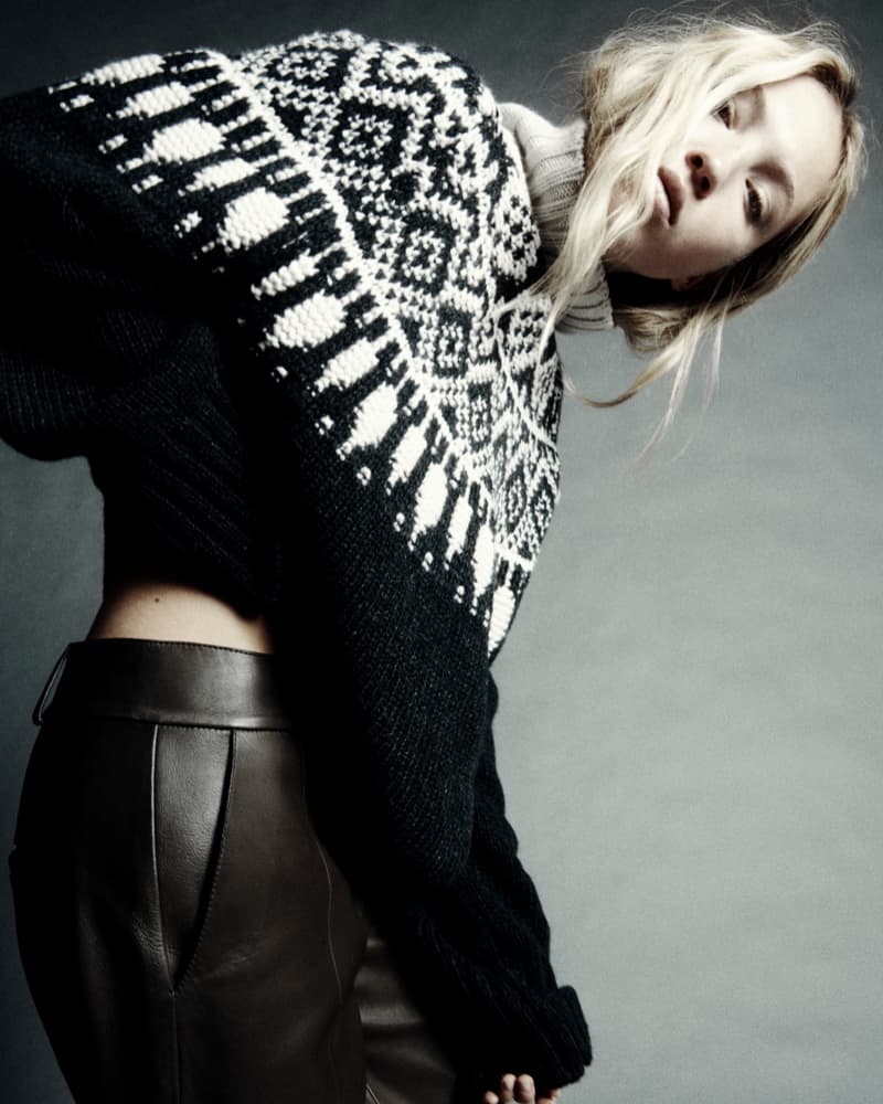 A stylish woman with blonde hair leans forward, showcasing a black and white patterned sweater and brown leather pants against a neutral background.