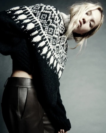 A stylish woman with blonde hair leans forward, showcasing a black and white patterned sweater and brown leather pants against a neutral background.