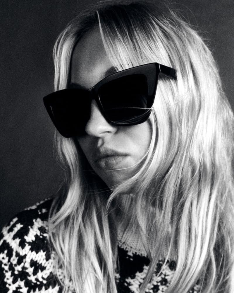 Black and white portrait of a woman wearing black cat-eye sunglasses and a patterned sweater, her blonde hair cascading down.