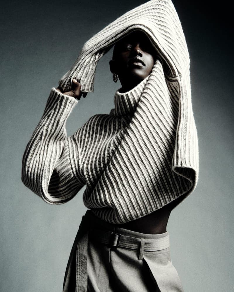 Fashion shot of an African-American woman wearing a white oversized sweater and gray skirt posing in front of a gray studio backdrop.