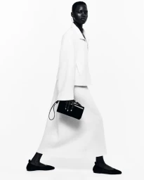 Model in white jacket and matching skirt holding a black clutch against a plain background