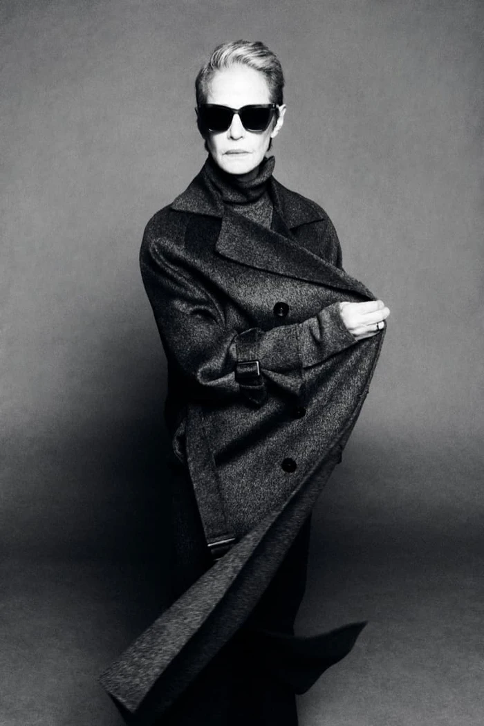 Charlotte Rampling wearing dark sunglasses and a draped charcoal wool coat over a turtleneck in black and white.