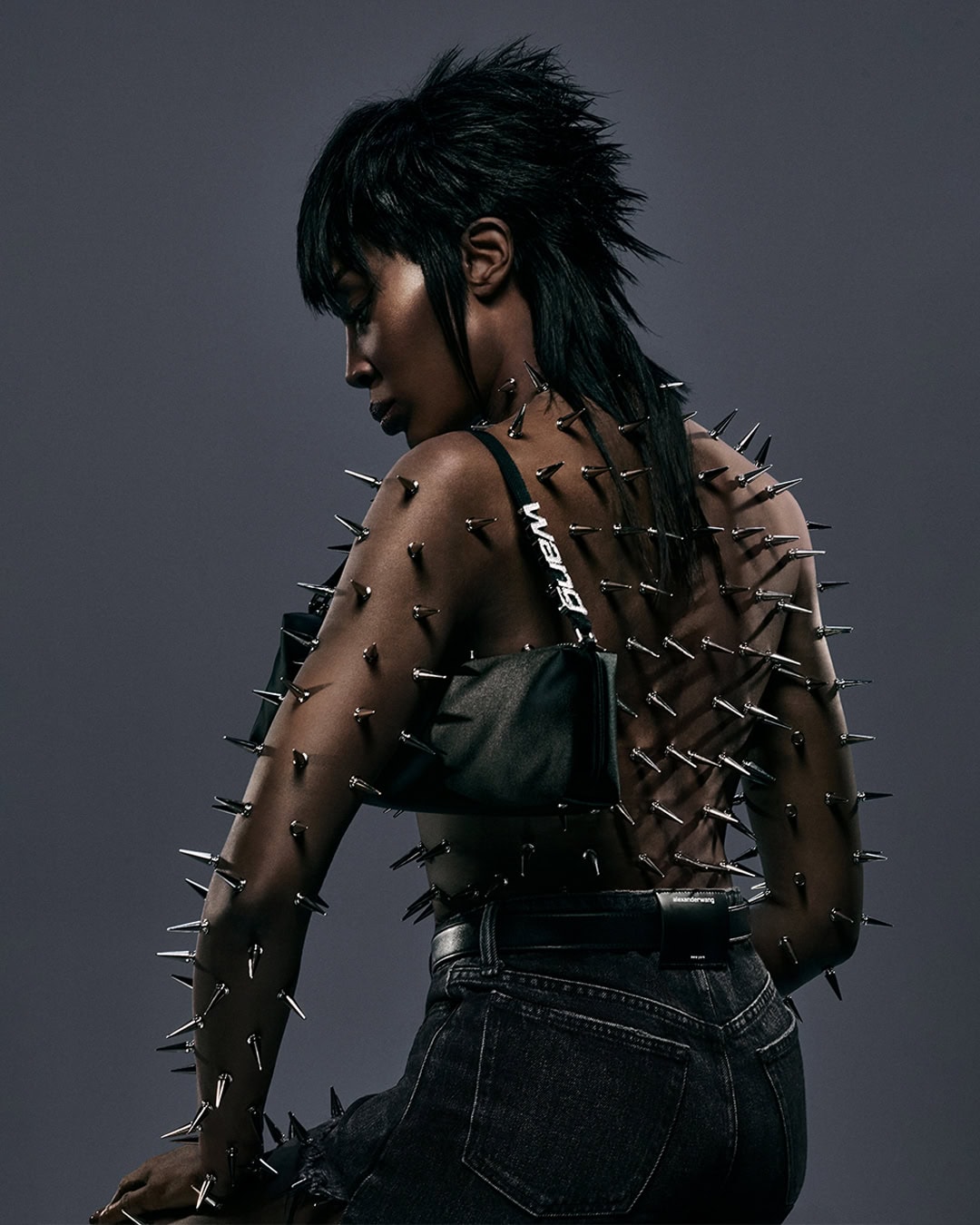 Fashionable woman with dark hair and exposed back decorated with metal spikes, wearing dark jeans and a black shoulder bag. 