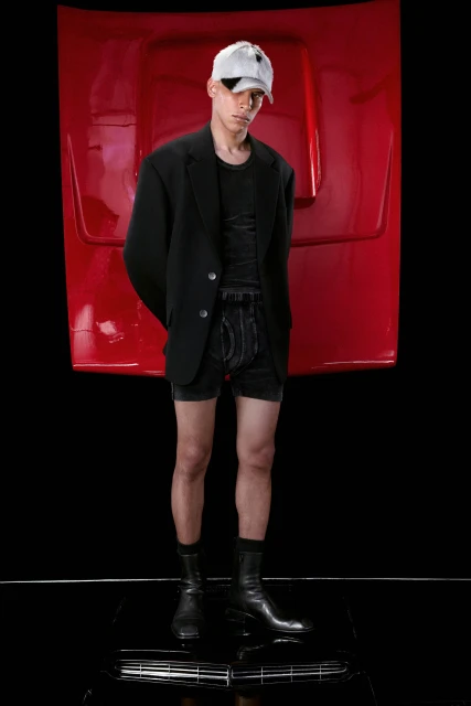 Young adult man in black blazer, tee, and shorts posing in a studio with a red car hood backdrop and shiny black floor.