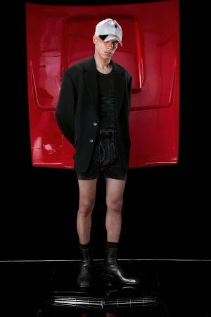 Young adult man in black blazer, tee, and shorts posing in a studio with a red car hood backdrop and shiny black floor.