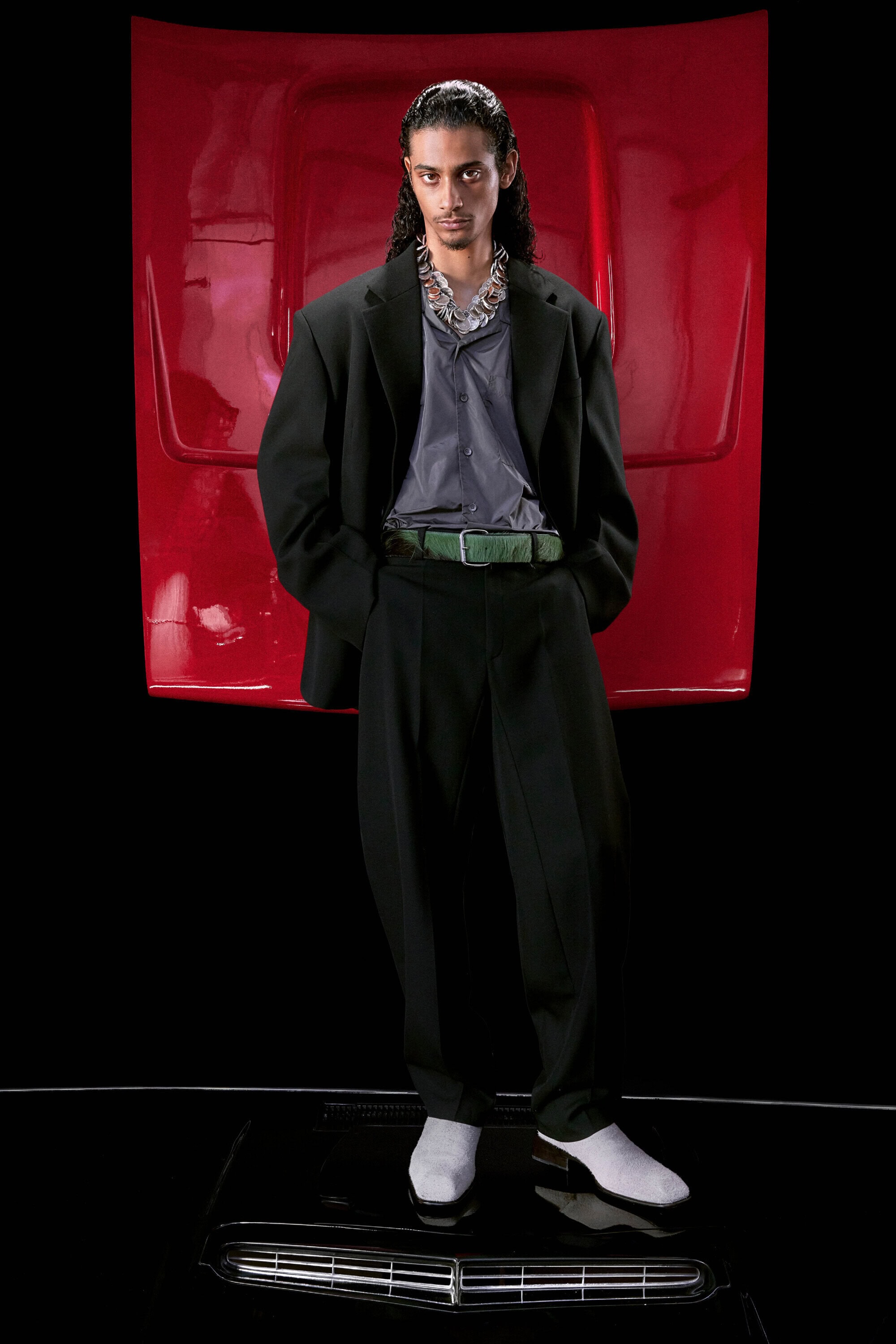 Man poses in oversized suit with car hood backdrop. Dark hair, statement necklace, shiny black boots, retro style, dramatic lighting.