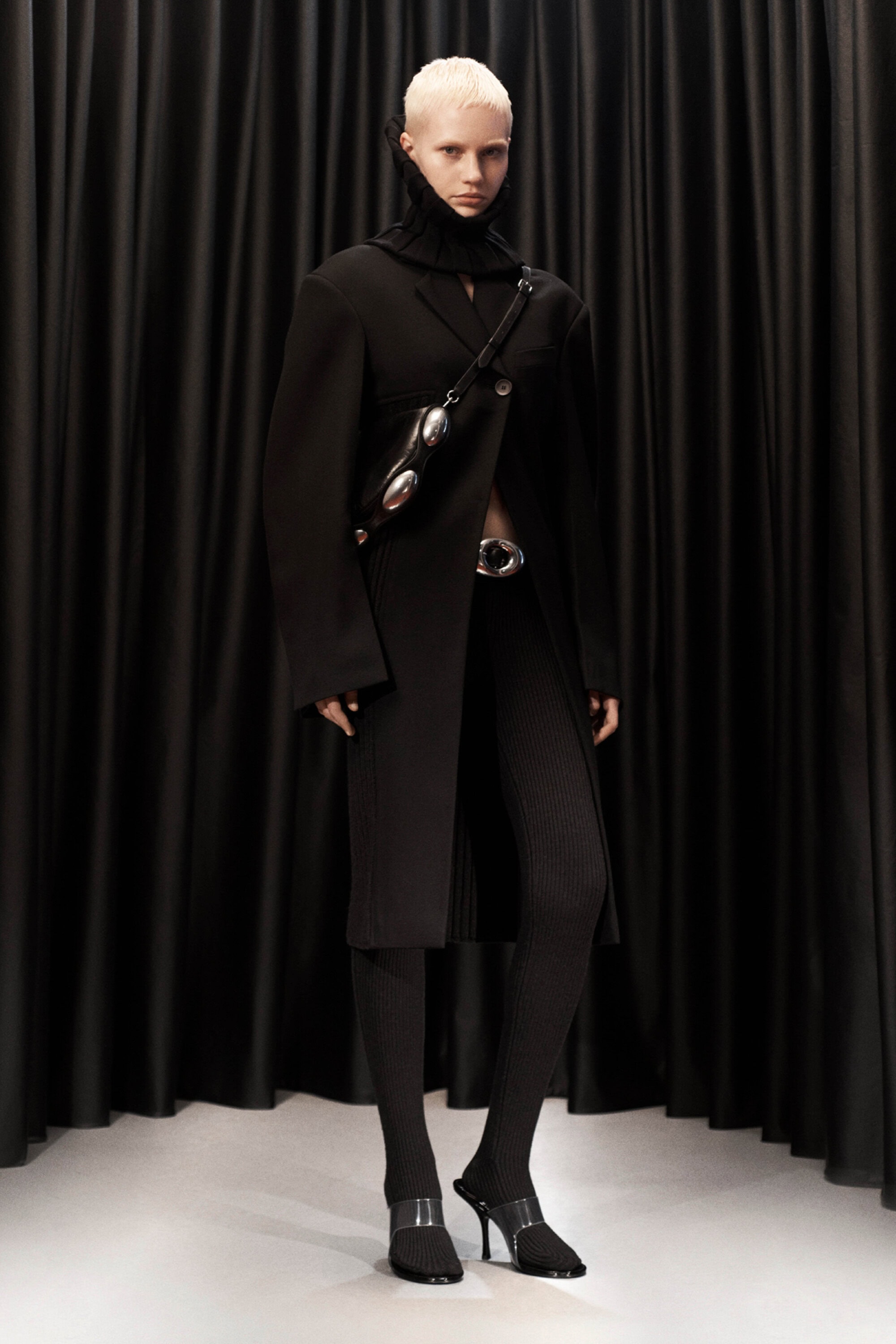 A light-skinned fashion model poses in a long black coat, textured leggings, and high heels against a dark curtain backdrop in a studio setting.
