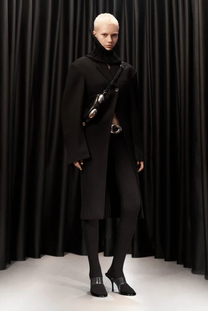 A light-skinned fashion model poses in a long black coat, textured leggings, and high heels against a dark curtain backdrop in a studio setting.