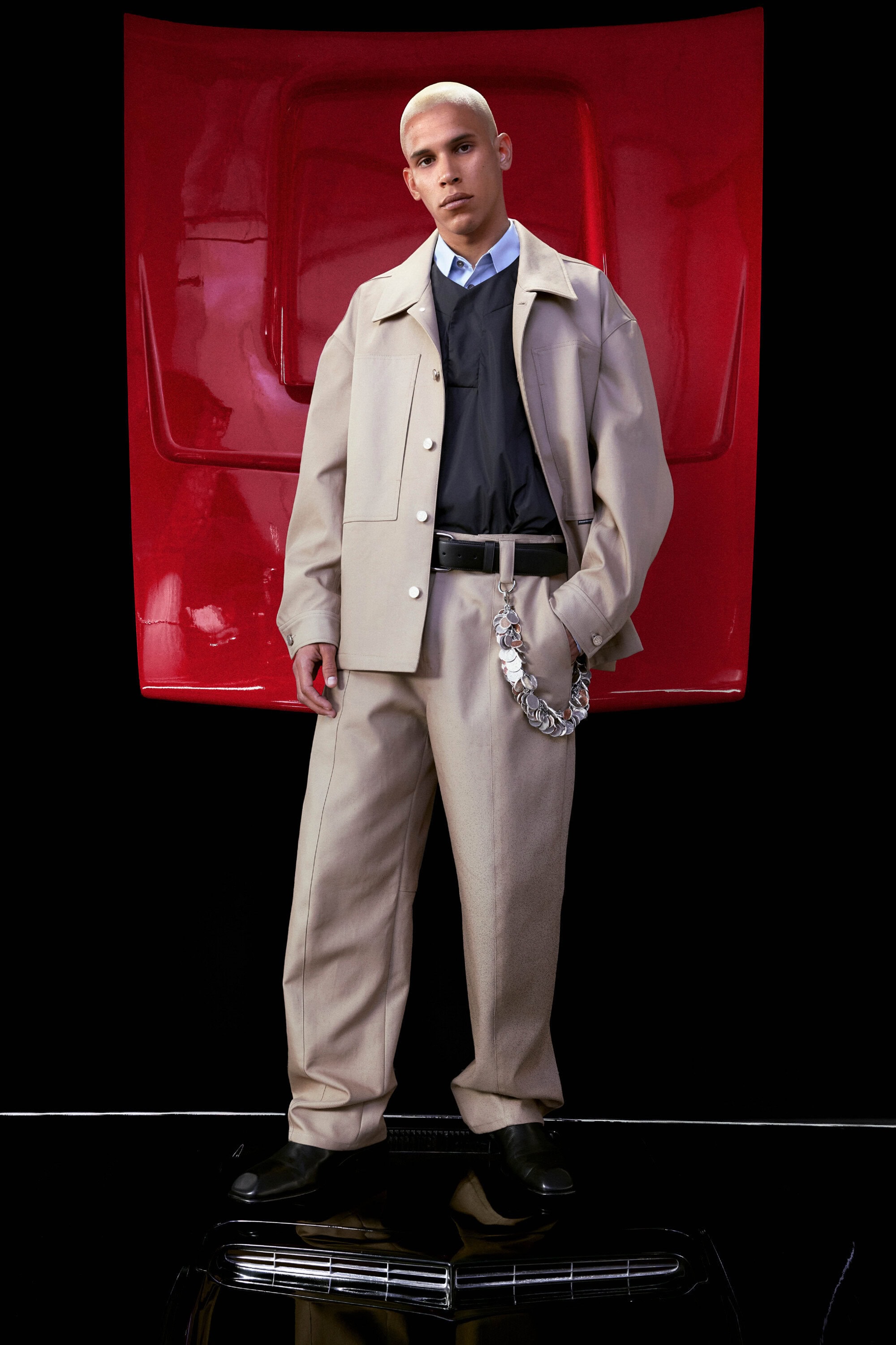 Male fashion model in a beige jacket and pants poses in a studio with a red car hood prop and a shiny black floor.