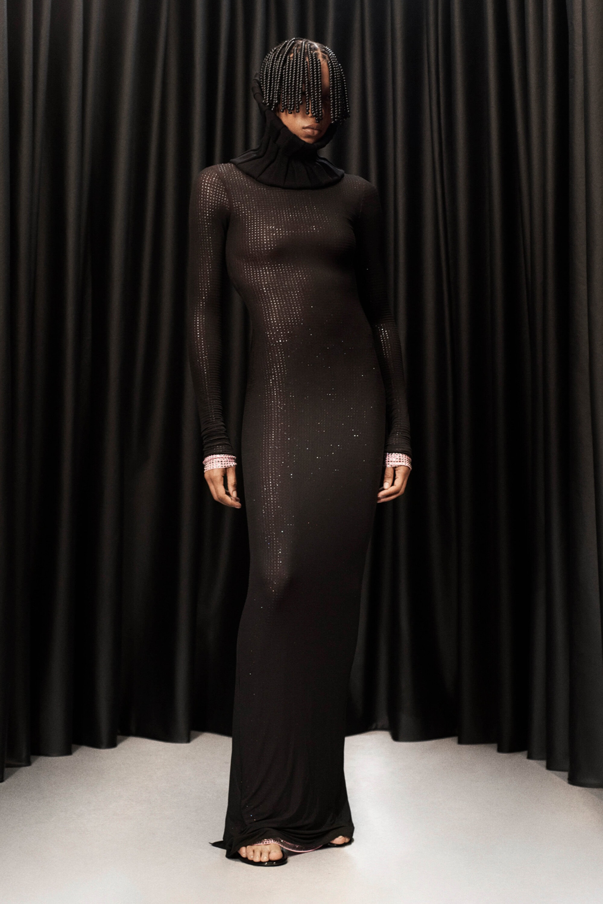 A model stands against a dark curtain backdrop, wearing a long black dress with long sleeves and a turtleneck collar.