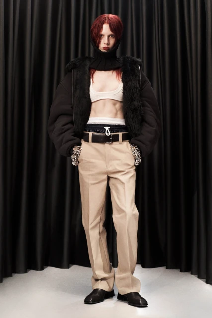 Full-length portrait of a young adult woman with red hair wearing a black jacket, top and neutral trousers against black drapes.