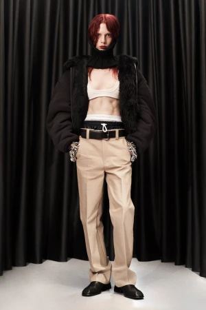 Full-length portrait of a young adult woman with red hair wearing a black jacket, top and neutral trousers against black drapes.