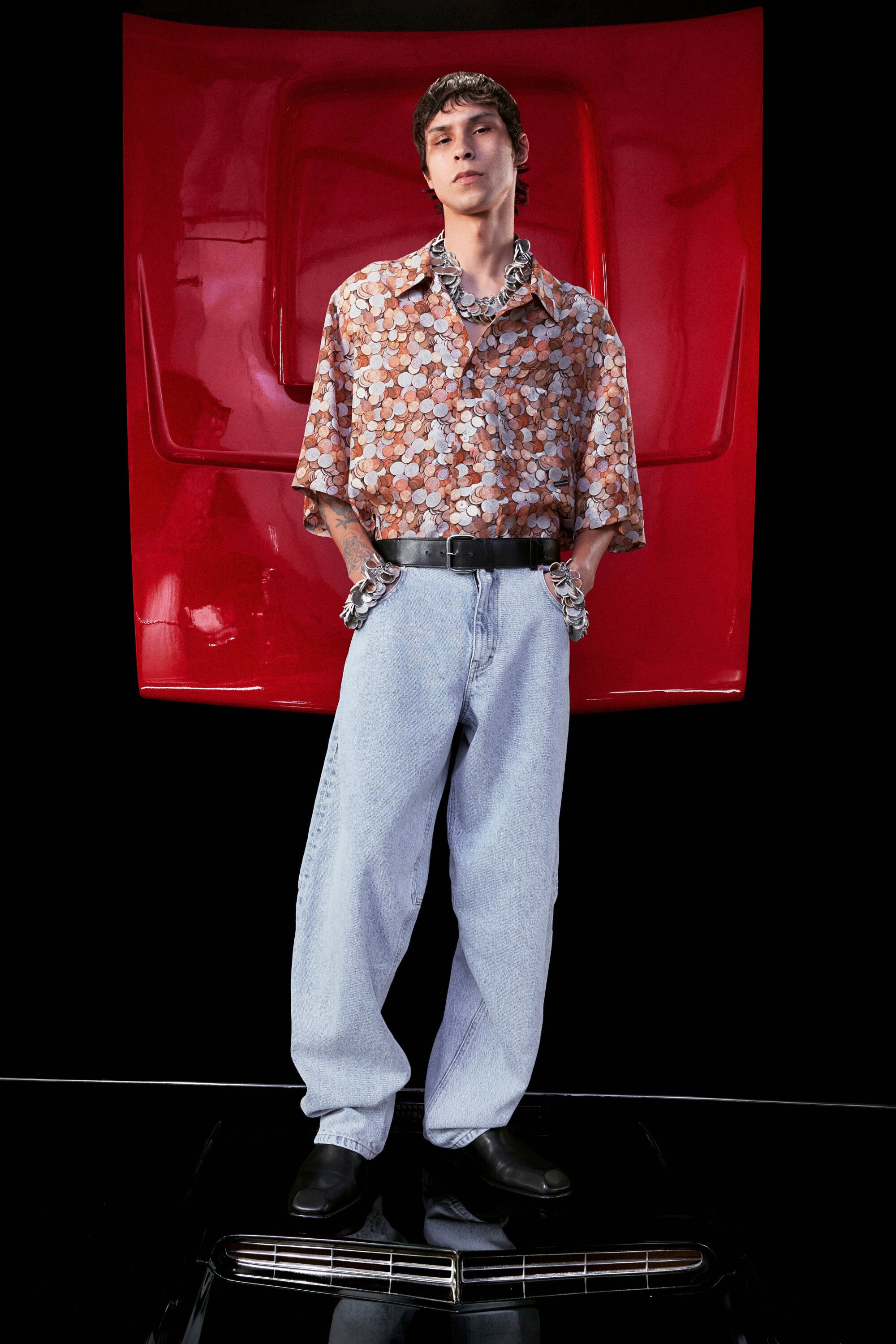 A young adult man posing in a floral short sleeve shirt and light blue jeans in front of a red car hood in a studio setting.
