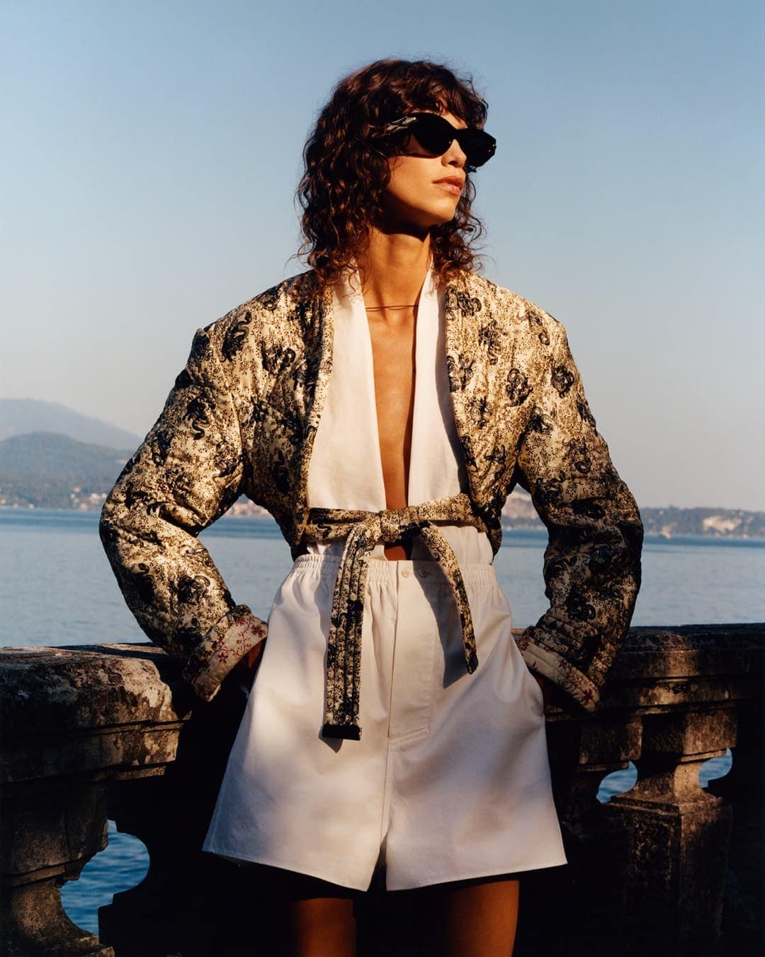 Fashion shot of a woman in a patterned jacket and white shorts posing outdoors with water and distant mountains in the background.