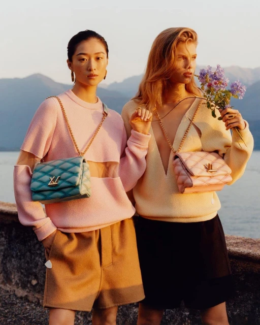 Two women model designer quilted bags with gold chain straps, wearing coordinated neutral sweaters, outside with a lake and mountains.