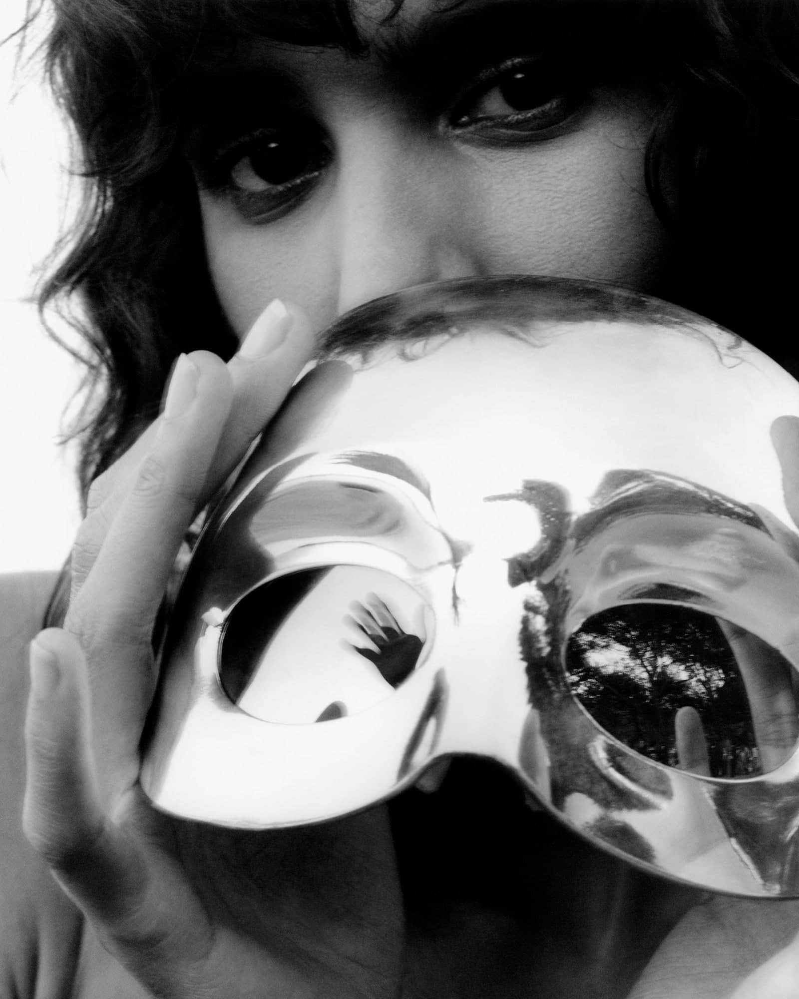 Portrait of a person holding a shiny mask in front of their face, obscuring the lower half, with a glimpse of eyes above.