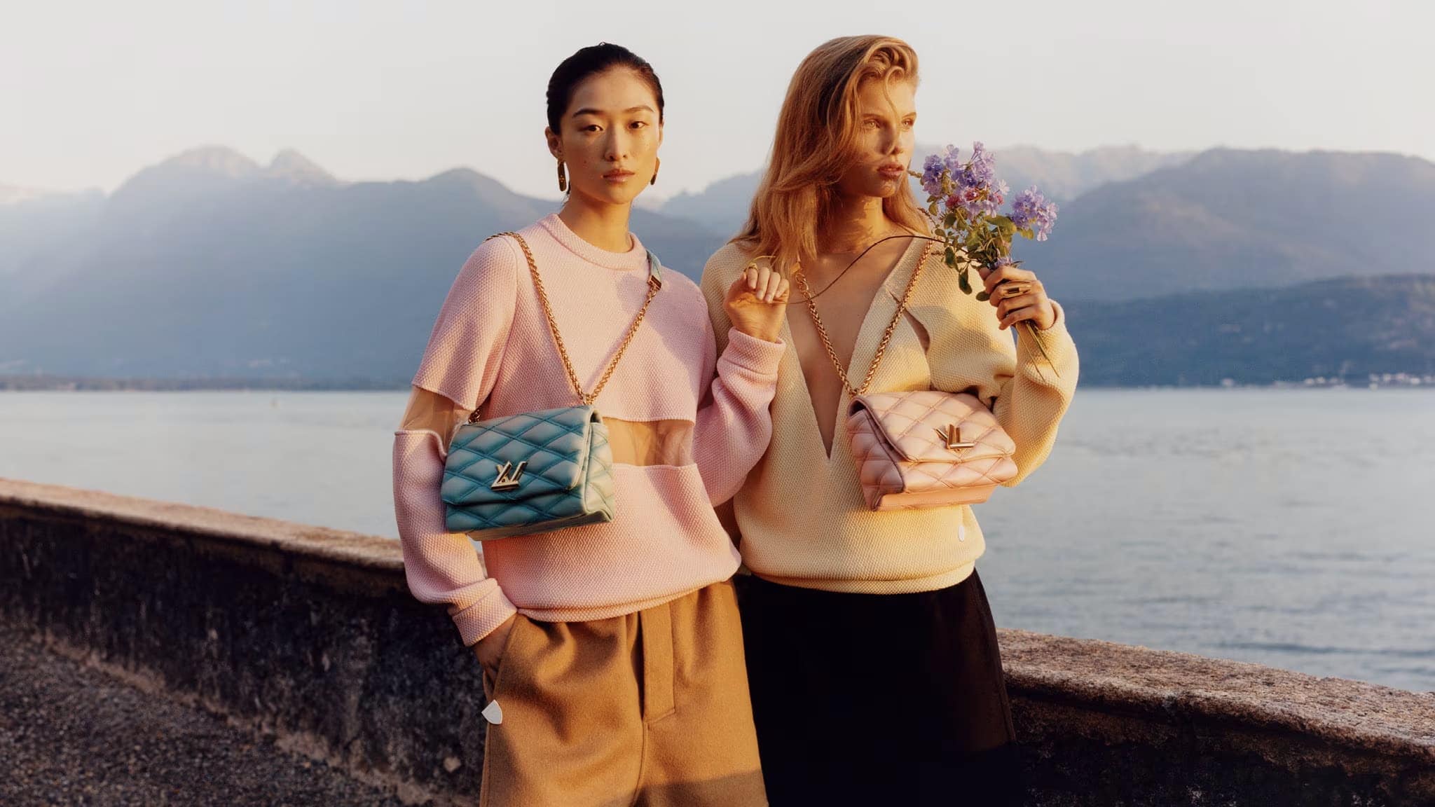 Two women pose near a lake with mountains in the background, showcasing fashionable handbags with a knitted sweater and neutral pants.