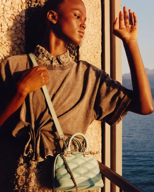 Woman in grey shirt and patterned skirt carrying a blue purse with a gold chain standing near a concrete structure by the water.