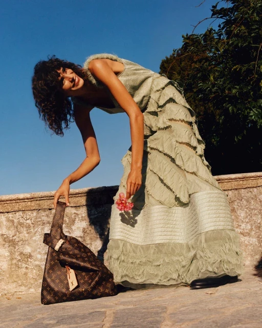 Brunette fashion model poses outdoors in a tiered green dress and a Louis Vuitton bag with flowers near a stone wall.