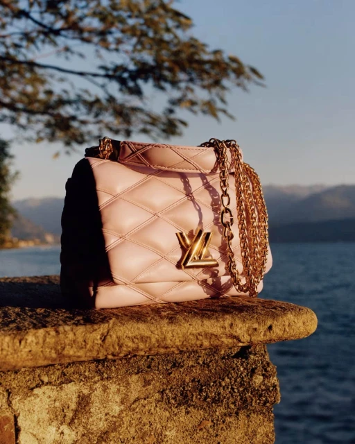 Designer quilted pink leather shoulder bag with a gold chain strap and logo on a stone ledge with a lake and mountains in the background.