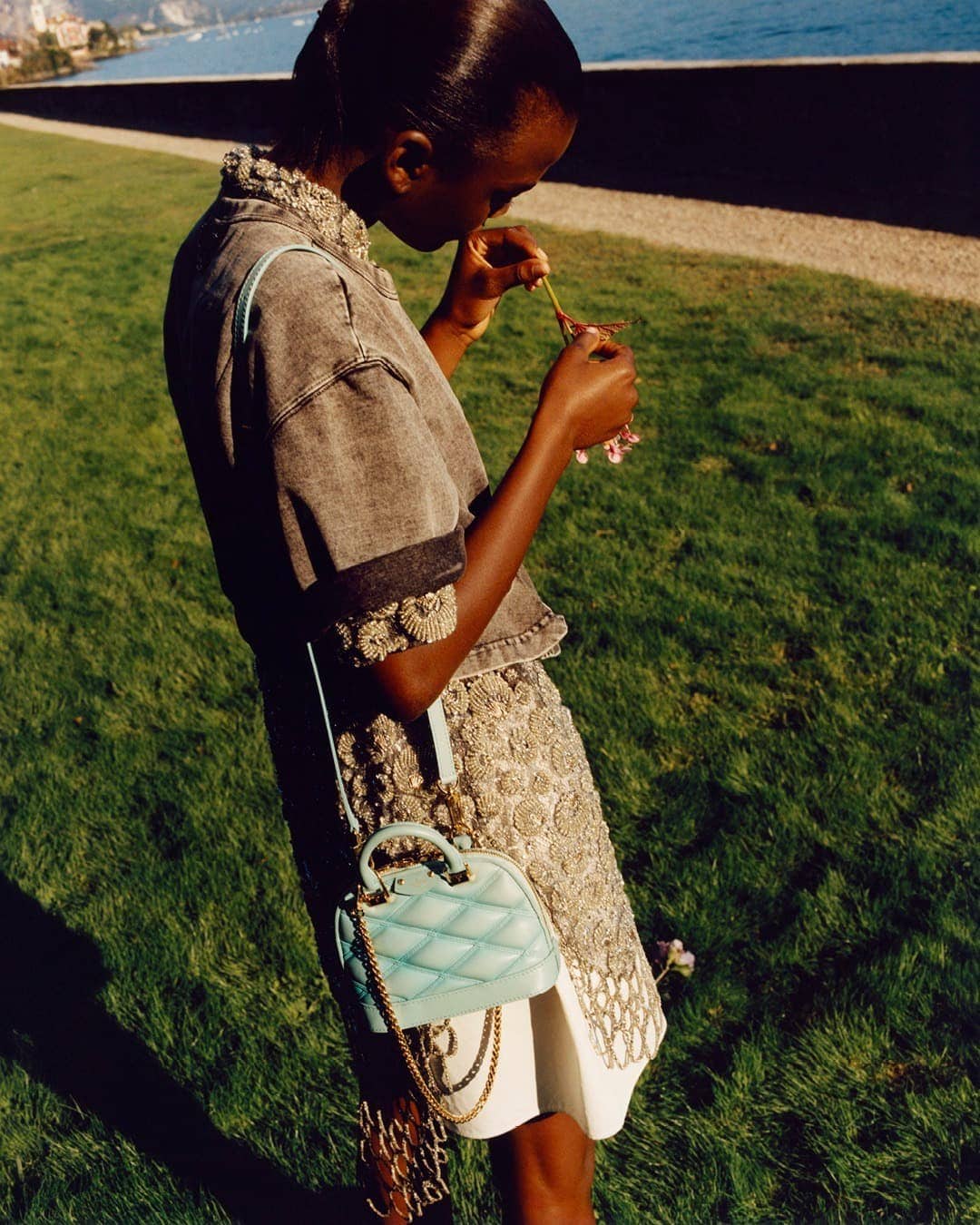 Dark-skinned woman in a grey jacket and ornate skirt stands on grass, smelling a flower, with a light blue purse and lake backdrop.