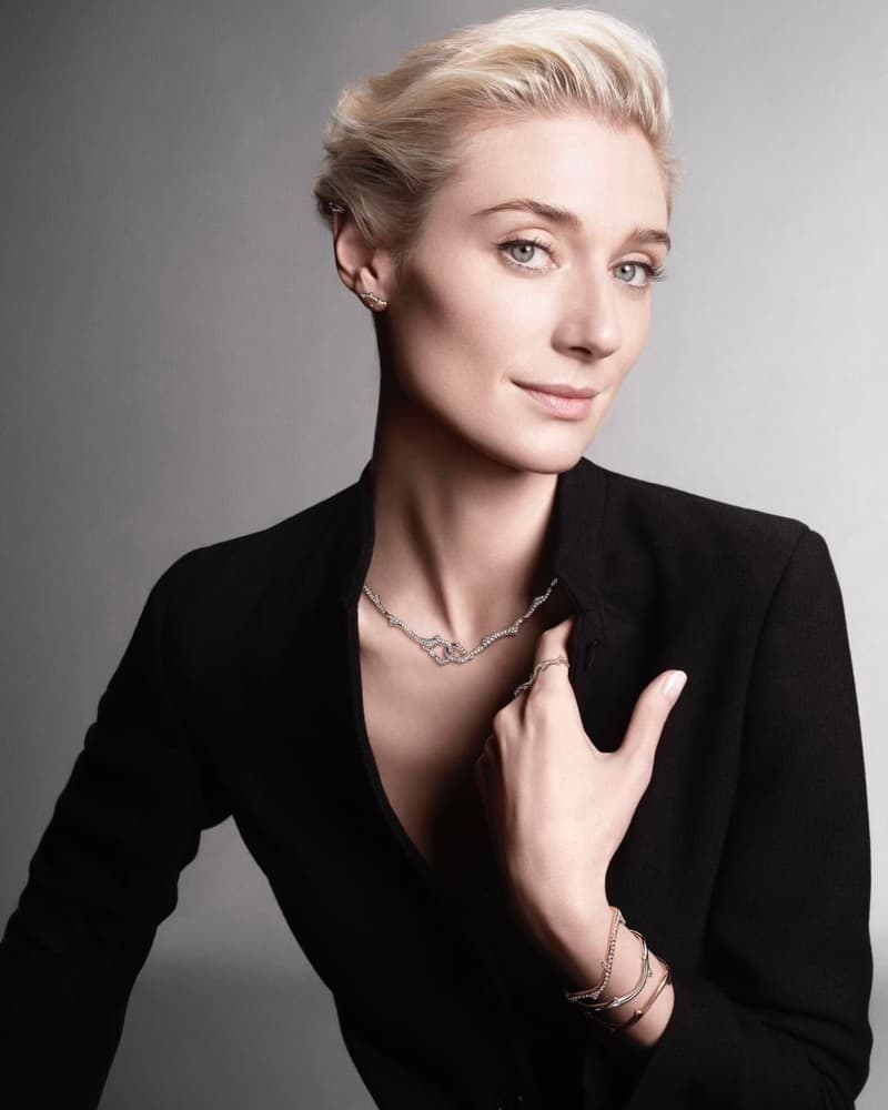 Elegant woman with short blonde hair poses in a black jacket with jewelry, in a studio shot with a neutral gray backdrop.
