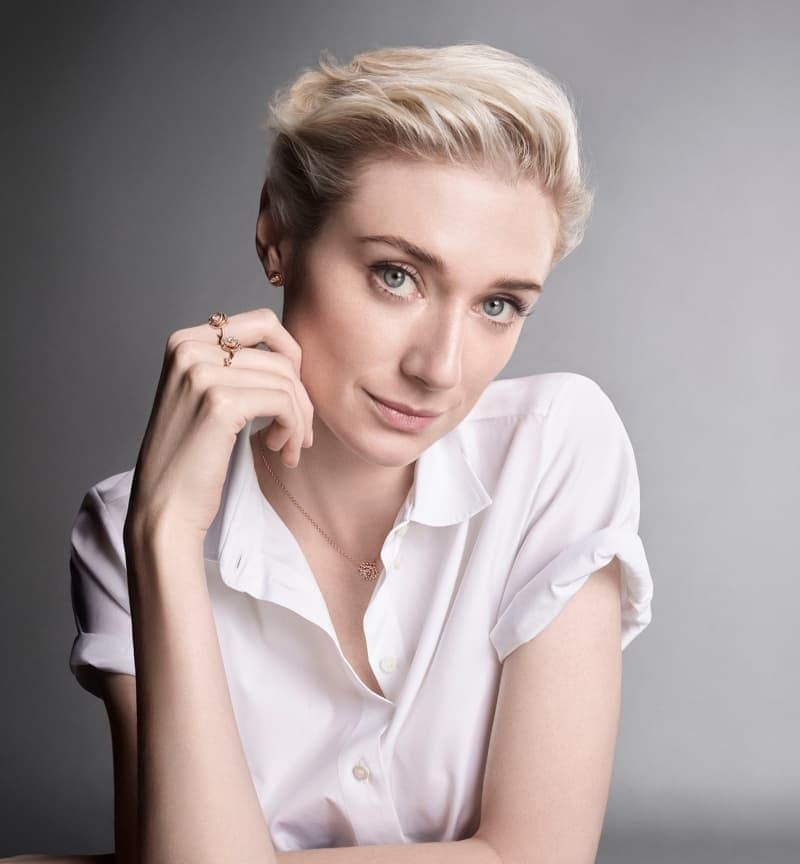 Elizabeth Debicki wearing fine gold jewelry and a short-sleeved white button-down shirt against a grey studio backdrop.