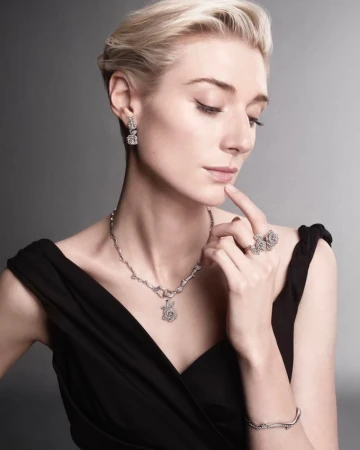 Woman with short blonde hair wearing elegant black dress and diamond jewelry, including necklace, earrings, ring, and bracelet, on gray backdrop.