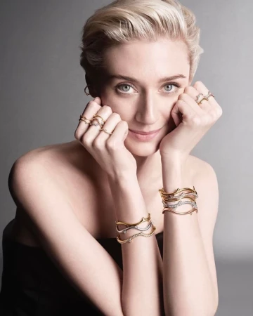 Close-up of an adult woman wearing multiple gold rings and bracelets posing in front of a grey background in a beauty shot.
