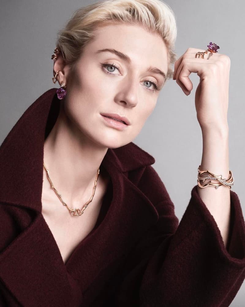 Close-up of an adult woman with blonde hair and fair skin wearing a maroon coat and gold jewelry against a grey backdrop.