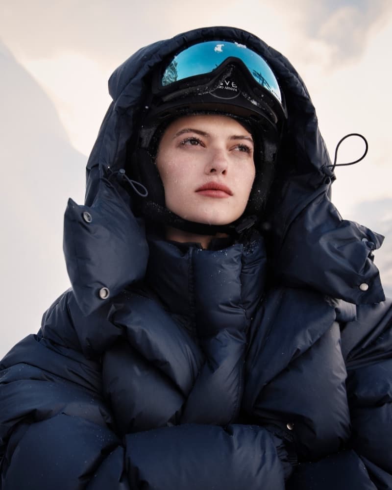 Woman in a black ski helmet and goggles, wearing a puffy navy blue jacket, looks off into the distance against a blurred background.