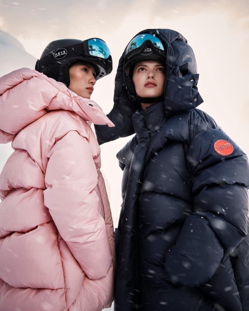 Two women wearing ski helmets and large puffer jackets stand in a snowy landscape. One jacket is pink and the other is navy blue.