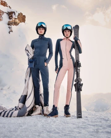 Two women in ski suits and helmets pose with skis in a snowy mountain setting, showcasing winter fashion apparel and outdoor adventure.