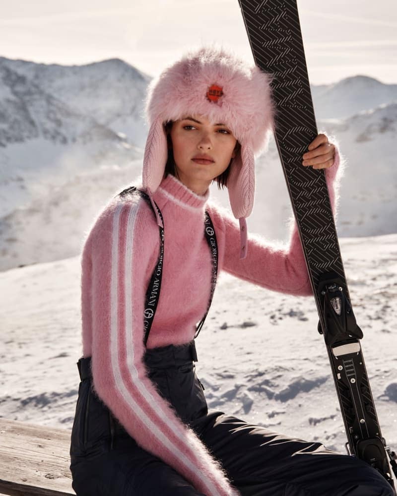 Woman in pink sweater and fur hat sitting in snow holding black skis with mountain backdrop on sunny day in a fashion editorial shoot.