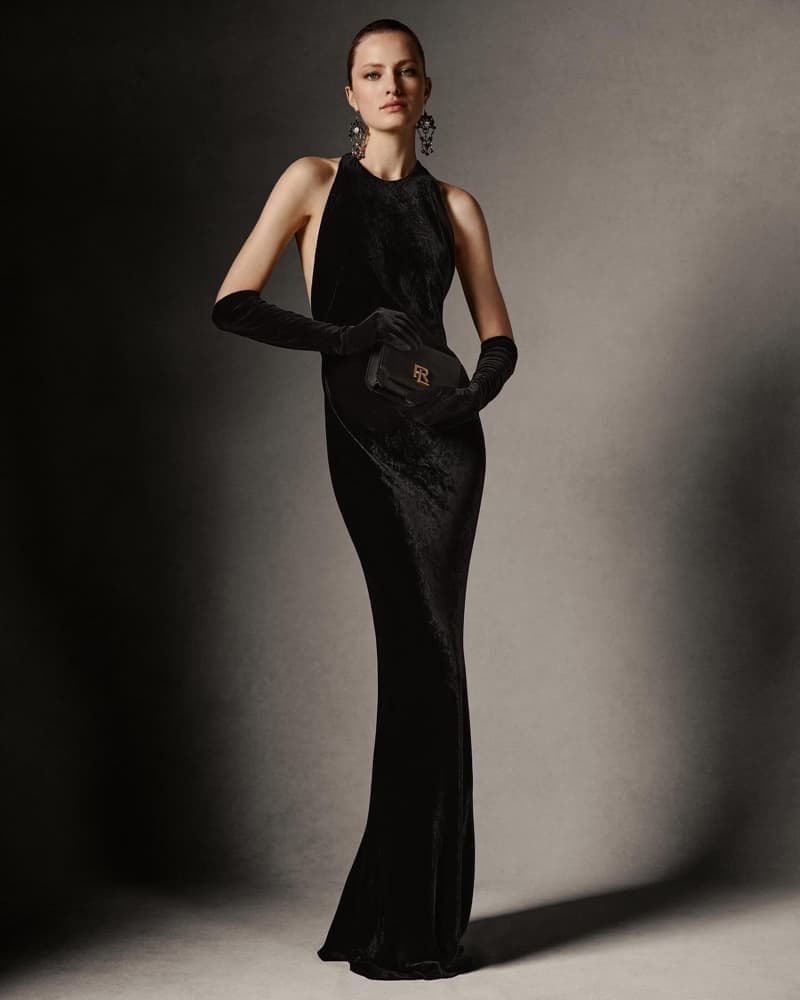 A woman in a black halter-style gown and long gloves holds a black clutch with logo against a gradient dark gray background.