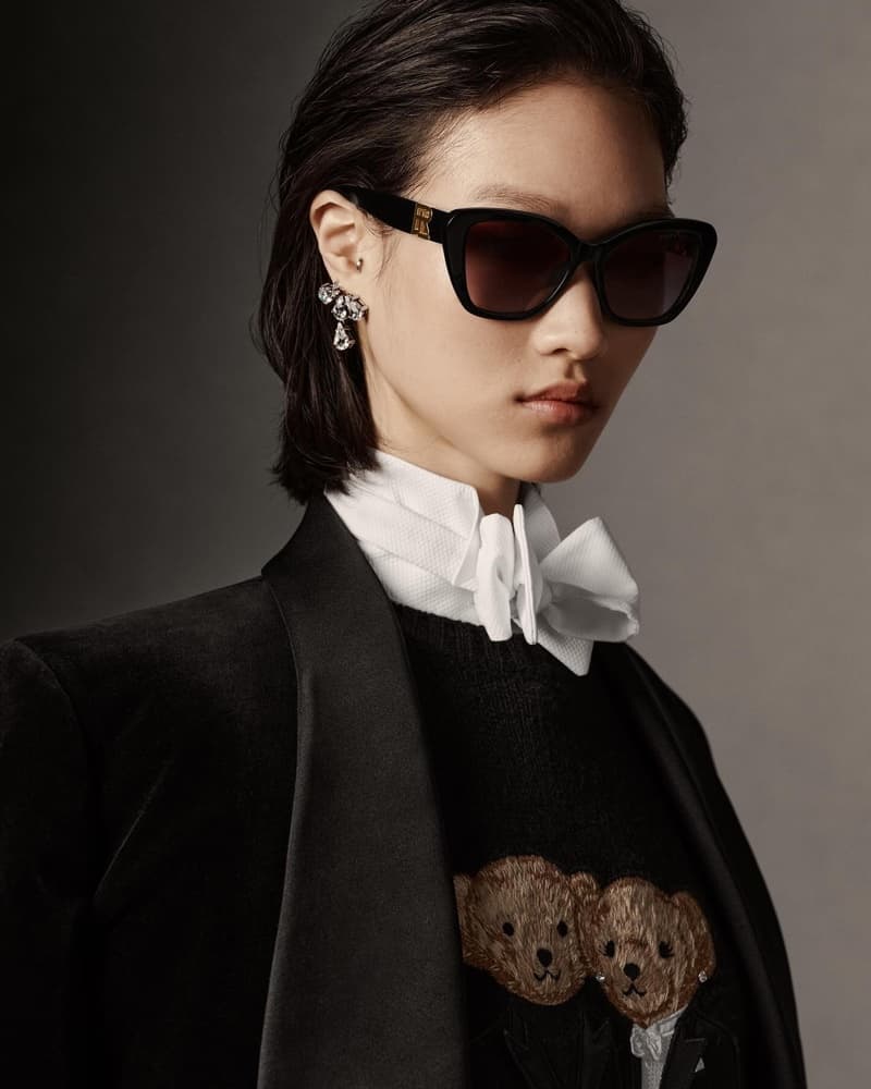 Headshot of a model wearing dark sunglasses, a white ruffled shirt, a black blazer, and statement earrings against a gray background.